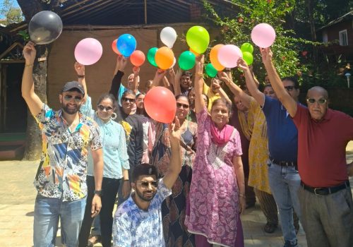 A diverse group of people of all ages smiling and holding colorful balloons during a sunny outdoor celebration at a nature resort.